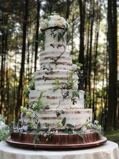 a wedding cake with white flowers and greenery on the top is surrounded by trees