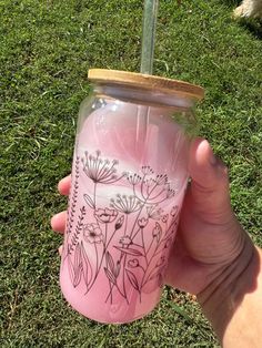 a hand holding a pink mason jar with flowers painted on the side and a straw lid