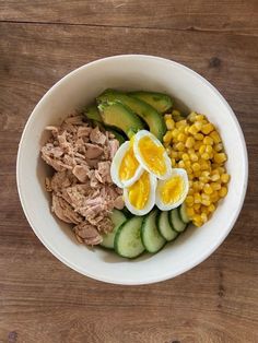 a white bowl filled with meat, boiled eggs and cucumbers on top of a wooden table