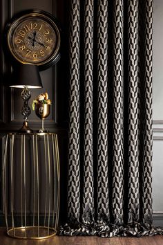 a clock sitting on top of a gold table next to a window covered in black and white curtains