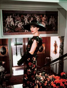 a woman in a black hat and dress stands on the stairs next to some flowers