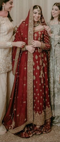 three women standing next to each other in dresses and veils, one wearing a red bridal gown