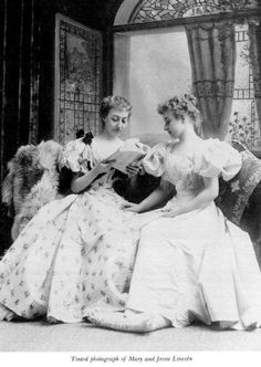 an old photo of two women in dresses sitting on a couch and one is reading a book