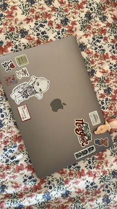 an apple laptop with stickers on it sitting on a flowered surface, next to a doll