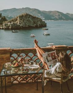 a woman reading a magazine while sitting at a table with a view of the ocean