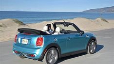 a small blue convertible car driving down the road near the ocean and sand dunes in front of it