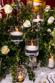 candles are lit in glass vases with greenery and roses on the table top