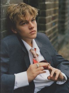 a young man in a suit leaning against a brick wall with his hand on his tie