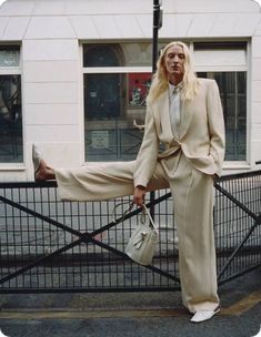 a woman in a suit leaning on a fence with her legs crossed and holding a handbag