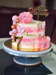 a pink and gold birthday cake with flowers on top is sitting on a glass table