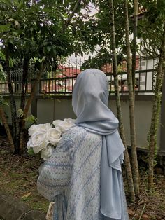 a woman wearing a blue shawl and holding white flowers in front of some trees