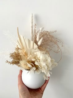 a hand holding a white vase with dried flowers and feathers in it, against a white wall