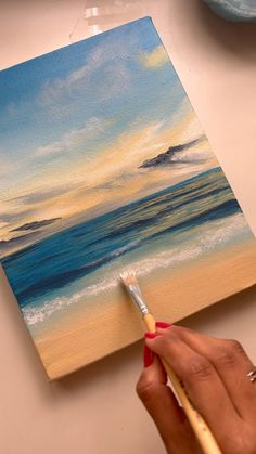 a person is holding a paintbrush in their hand and painting a sunset on the beach