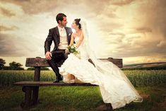 a bride and groom sitting on a bench in the middle of a field under a cloudy sky