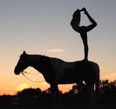 a woman standing on top of a horse in the middle of a field at sunset