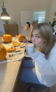 a woman sitting at a table drinking from a cup with pumpkins on the table behind her