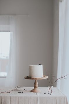 a small white cake sitting on top of a wooden stand next to a window with sheer curtains