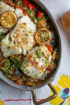 a pan filled with fish and vegetables on top of a white table cloth next to bread