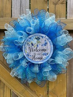 a blue mesh wreath with the words welcome to our home written on it in front of a wooden fence