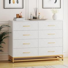 a white dresser with two pictures on the wall above it and a potted plant next to it