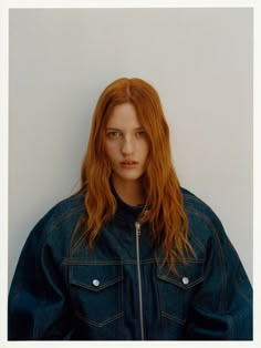 a woman with long red hair wearing a denim jacket and standing in front of a white wall