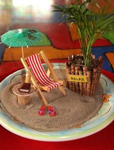 a miniature beach scene with an umbrella, chair and potted plant in the sand