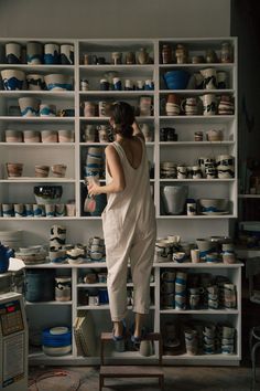 a woman standing on top of a wooden stool in front of shelves filled with pottery