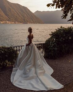 a woman in a wedding dress looking out over the water with mountains in the background