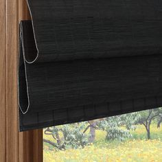 a black roman blind hanging on the side of a window in front of trees and yellow flowers