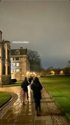 two people walking down a walkway in front of a large building at night with lights on