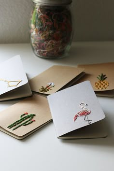 four notebooks with embroidered designs on them sitting on a table next to a jar