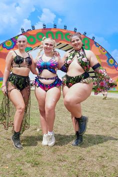 Three festival-goers in detailed and colorful outfits with floral motifs pose together in front of a multicolored arch. They are smiling and standing on a grassy field under a sunny sky.