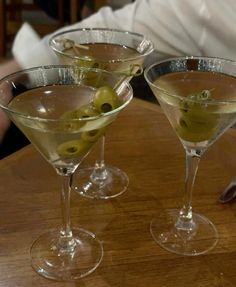 three martini glasses with olives in them on a wooden table next to a man