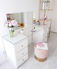 a white desk topped with a mirror and pink stool next to a shelf filled with flowers