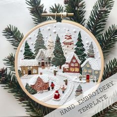 a hand embroidered christmas ornament with houses and trees in the snow, surrounded by pine needles