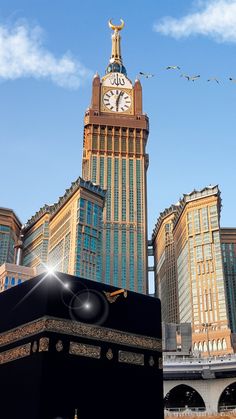 a large black object with a clock on top in front of some buildings and birds flying overhead