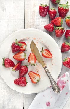 strawberries on a white plate with a knife and fork next to it, surrounded by other strawberries