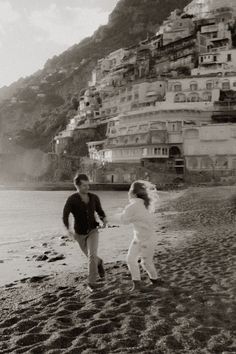 a man and woman walking on the beach with buildings in the background, black and white photo