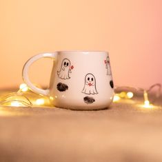 a white coffee cup with ghost faces on it sitting on a table next to some string lights