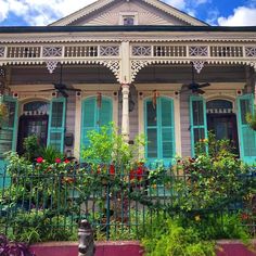 an old house with green shutters and flowers on the front porch, surrounded by greenery