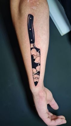 a knife tattoo on the left forearm and hand with flowers around it, in black ink