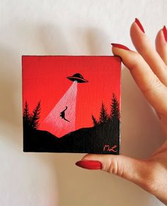 a hand holding up a small square painting with a red sky and trees in the background