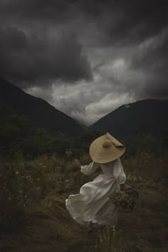 a woman in a white dress and straw hat walking through a field with mountains in the background