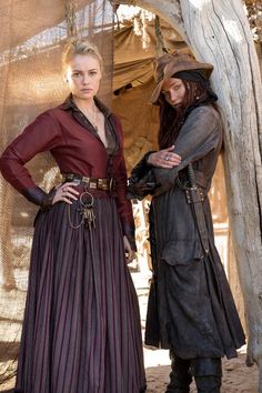 two women dressed in medieval clothing standing next to each other on a dirt ground near a tree