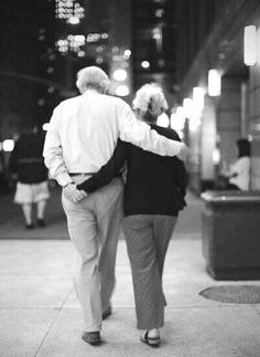 an older couple walking down the sidewalk in front of a building at night, back to back