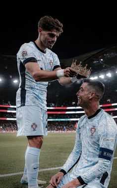 two soccer players are sitting on the field and one is holding a crown while the other holds his hand