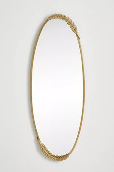 an oval shaped mirror with gold leaves on the rim and bottom, against a white wall