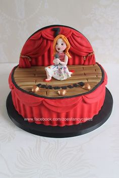 a cake decorated with a doll sitting on top of a wooden bench in front of a red curtain