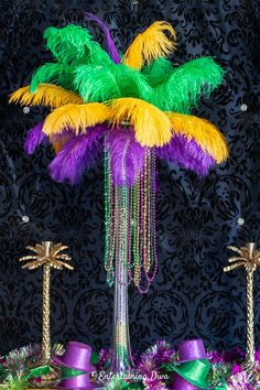 a table topped with purple, green and yellow feathers next to palm trees in front of a black wall