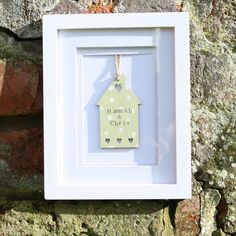 a white frame with a small house hanging from it's side next to a brick wall